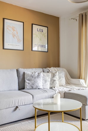 Een moderne woonkamer met een grijze bank, een witte salontafel met gouden poten, en twee ingelijste foto's aan de muur.