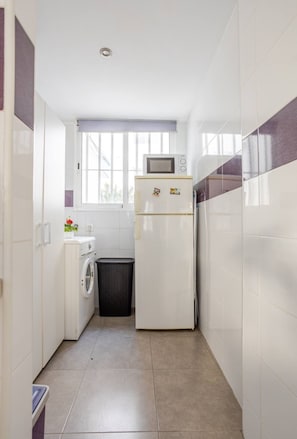 A compact kitchen with a washing machine, a small trash bin, and a refrigerator. - Apartamento inteiro a 3 minutos da Praia de Gandia (Gandia)