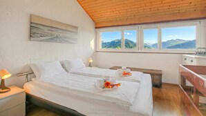 A bedroom with two beds, a wooden ceiling, a painting, a window with a mountain view, and a desk with a chair. - Angelina: Panoramic Lake View House on Monte Brè | Terrace & Balcony, Lugano, Switzerland (Lugano)