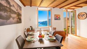 A dining area with a wooden table set for four, a painting of a mountain landscape, and a view of the outdoors through a window. - Angelina: Panoramic Lake View House on Monte Brè | Terrace & Balcony, Lugano, Switzerland (Lugano)