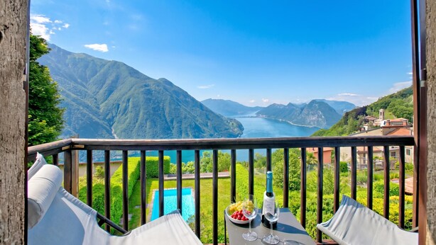 A balcony with a table, a bottle, and a glass of wine, overlooking a mountainous landscape and a body of water. - Angelina: Panoramic Lake View House on Monte Brè | Terrace & Balcony, Lugano, Switzerland (Lugano)