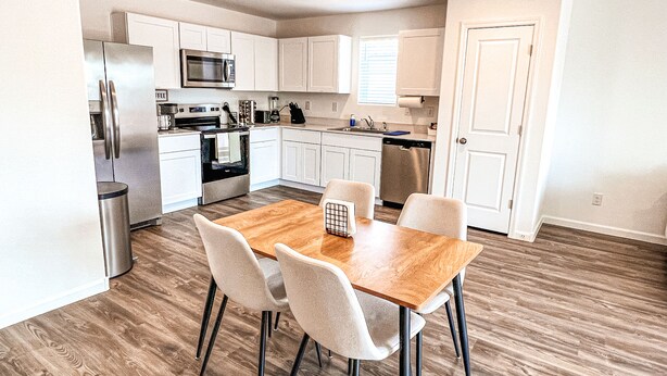 Una cocina moderna con armarios blancos, electrodomésticos de acero inoxidable y una mesa de comedor de madera con cuatro sillas.