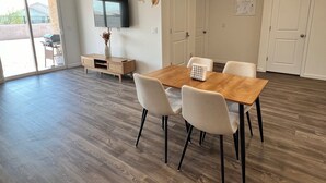 Un comedor con una mesa de madera y cuatro sillas, un televisor de pantalla plana montado en la pared, y un piso de vidrio corredizo que da a un patio exterior.