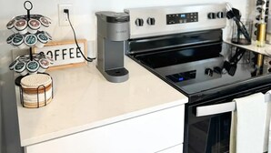 Una cocina con gabinetes blancos, microondas de acero inoxidable, cafetera Keurig y una estación de café con un letrero que dice "COFFEE".