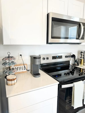 Una cucina con mobili bianchi, un forno a microonde in acciaio inossidabile, una macchina da caffè Keurig e un angolo caffè con un cartello che riporta la scritta 'COFFEE'.