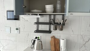 Eine Küche mit integriertem Mikrowellenherd, einem Wasserkocher, einem Holztisch zum Schneiden, einem Weinglas und einem Schrank mit Geschirr und Besteck.