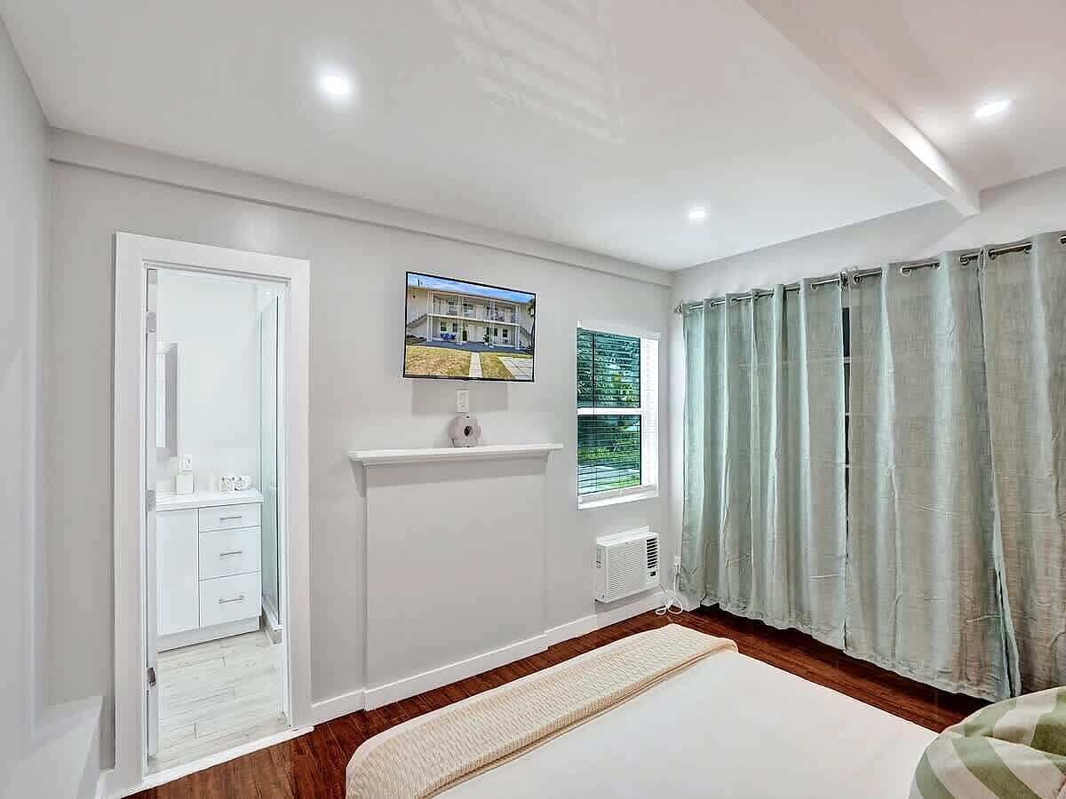 Une chambre à coucher avec un lit, une fenêtre avec des rideaux, un téléviseur fixé au mur et une porte donnant sur une salle de bain.