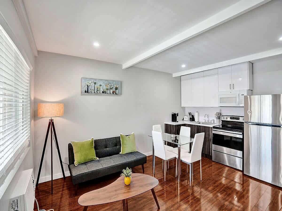 A modern living room with a sofa, dining table, chairs, a lamp, and a kitchen area with appliances.