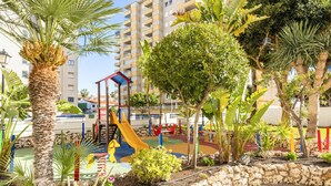 Un parque infantil con tobogán y estructuras para escalar, rodeado de palmeras y un muro de piedra.