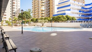 Zona de piscina con bancos de madera, piscina y edificios al fondo.