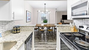 A modern kitchen with white cabinets, a granite countertop, a central island with stools, a stove, and a microwave. - Walk to Globe Life Field & AT&T Stadium - Free Parking - 2BR Sleeps 6 (Arlington)