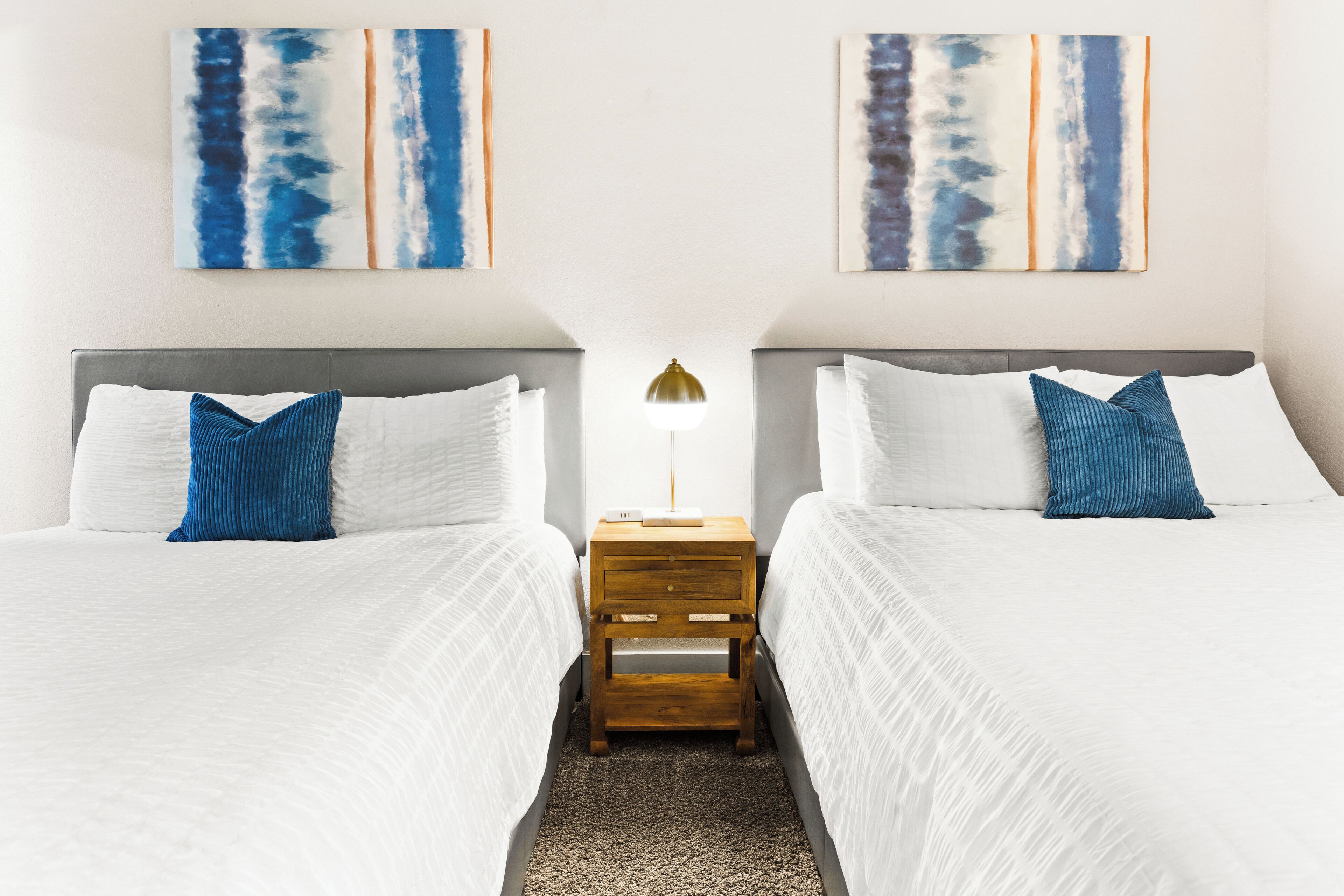 A hotel room with two beds, white bedding, a wooden nightstand, a lamp, and abstract art on the walls.
