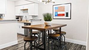 A modern kitchen with white cabinets, a wooden island with stools, a potted plant, and a framed American flag. - Walk to Globe Life Field & AT&T Stadium - Free Parking - 2BR Sleeps 6 (Arlington)