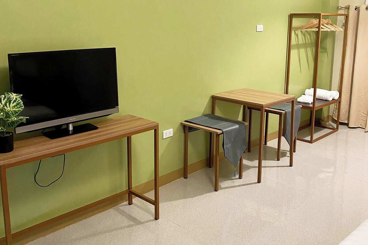 A room with a TV on a wooden table, a small stool, a wooden chair, and a clothing rack with hanging clothes.