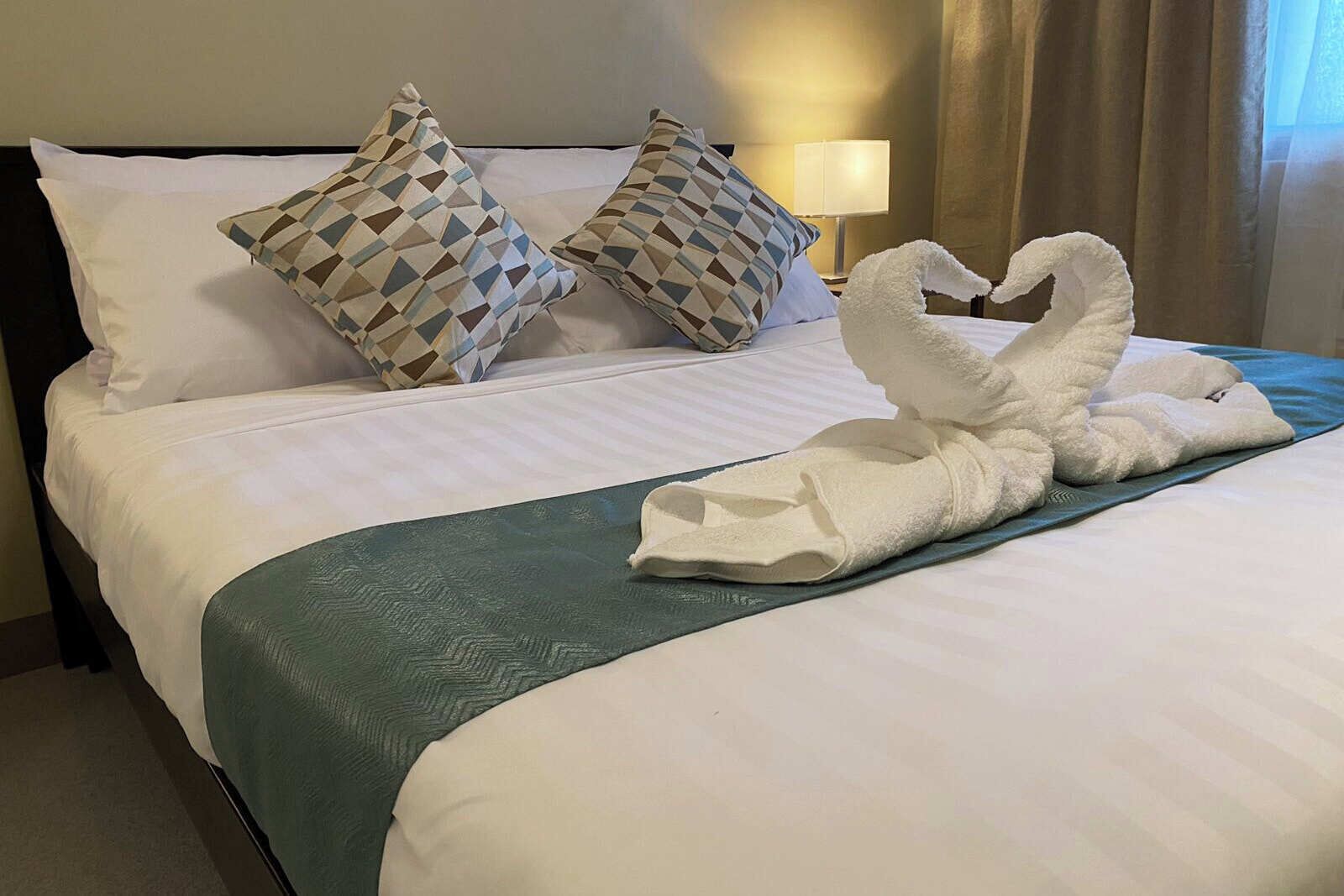 A neatly made bed with a green bedspread, two patterned pillows, and a towel shaped like swans.