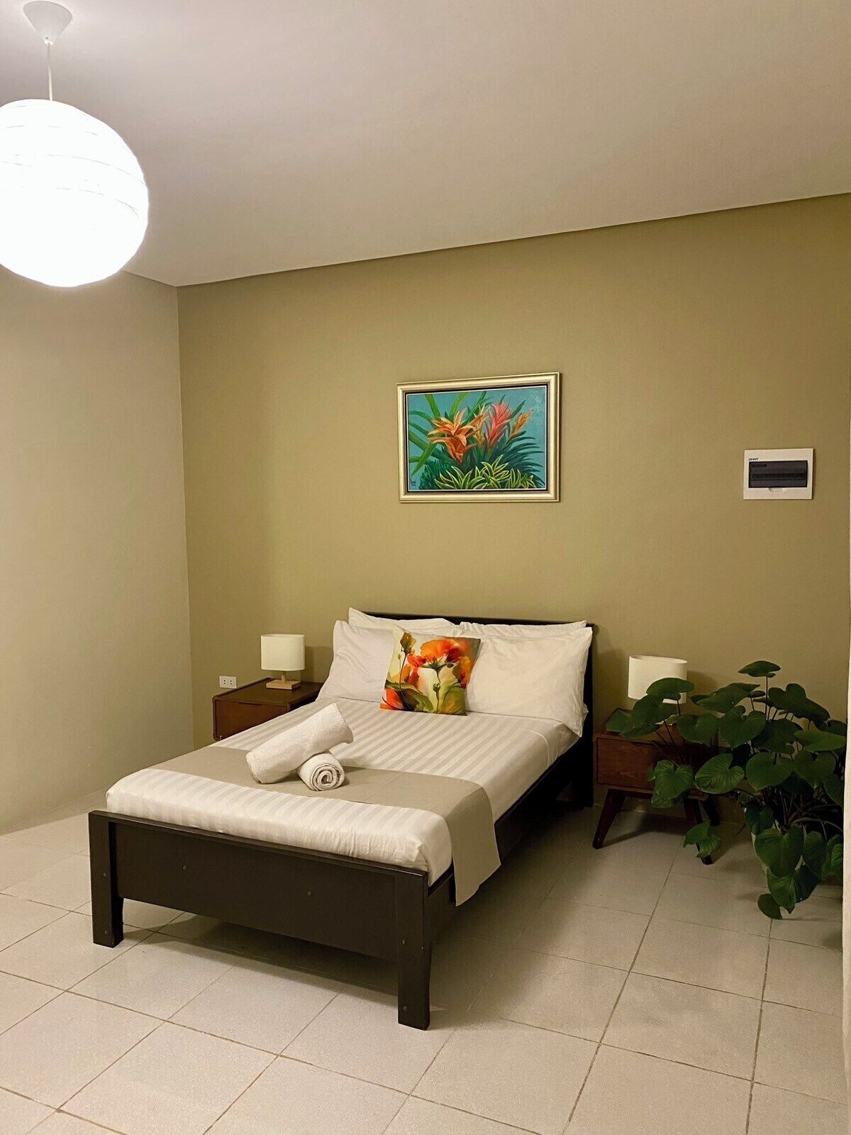 Una cama bien hecha con sábanas blancas, un solo almohadón y un cojín decorativo con motivos florales. Hay una mesita de noche con una lámpara, un aire acondicionado instalado en la pared y una planta en maceta a la derecha.