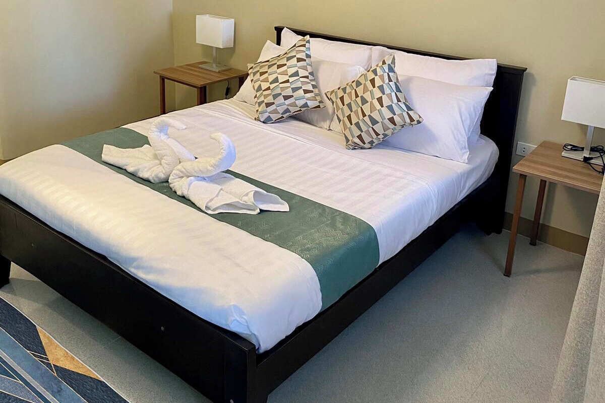 A neatly made bed with white linens, a green throw blanket, and two patterned pillows. There are two bedside tables with lamps, one on each side of the bed.