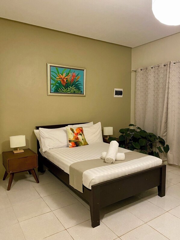 A neatly made bed with white linens, a nightstand with a lamp, a painting of flowers on the wall, and a potted plant beside the bed. - La Patria Baybay Tourist Inn powered by Cocotel (Baybay City)