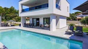 Ett modernt hus i två våningar med en swimmingpool, utomhusmöbler och utsikt över det omgivande landskapet.