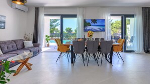 Ett modernt vardagsrum med en matplats, stora fönster och en platt-TV som visar en strandvy.