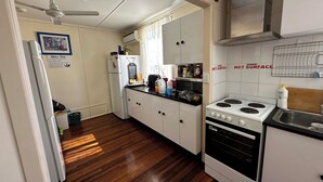 A compact kitchen with white cabinets, a microwave, a stove, and a sink. There is a ceiling fan, a framed picture on the wall, and a sign with kitchen rules. - Star Guesthouse (Proserpine)