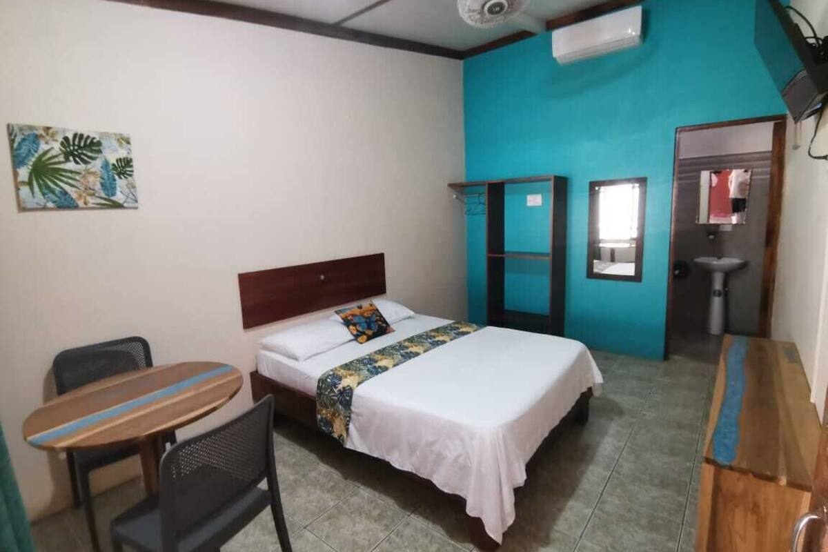 A room with a bed, a desk, a chair, a small table, a fan, and a sink.