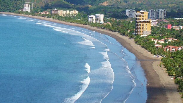 A coastal cityscape with a beach, buildings, and hills. - Hotel Cabinas Jacomar (Jacó)