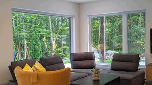 Una sala de estar moderna con un sofá, sillones y una mesa de centro. Amplias ventanas ofrecen vistas al verdor exterior.