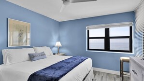 Ein Schlafzimmer mit Bett, Nachttisch, Lampe, Kommode, Fenster mit Jalousien und Deckenventilator.