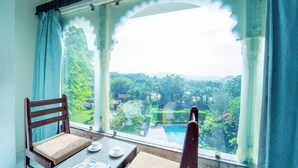 Une pièce avec vue sur une piscine et sur la verdure, une chaise en bois, une table avec un journal et une fenêtre avec des rideaux.