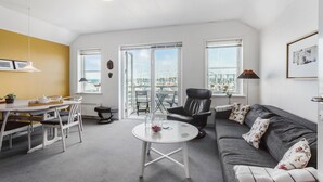Un salon moderne comprenant un canapé, une table à manger avec des chaises, une table basse et une vue sur une marina, visible à travers de grandes fenêtres.