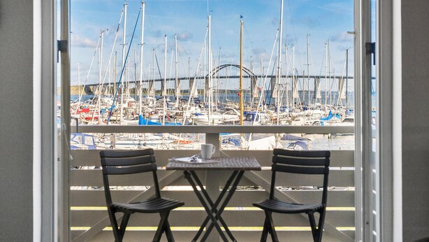 Un balcon avec deux chaises et une petite table, donnant sur une marina où se trouvent de nombreux voiliers, et avec un pont en arrière-plan.