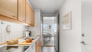 Une cuisine étroite avec des meubles en bois, un évier en acier inoxydable et un coin repas visible à travers une porte ouverte.