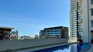 A rooftop swimming pool with a metal railing, surrounded by modern buildings and a clear sky. - Katara on Woods (Darwin)