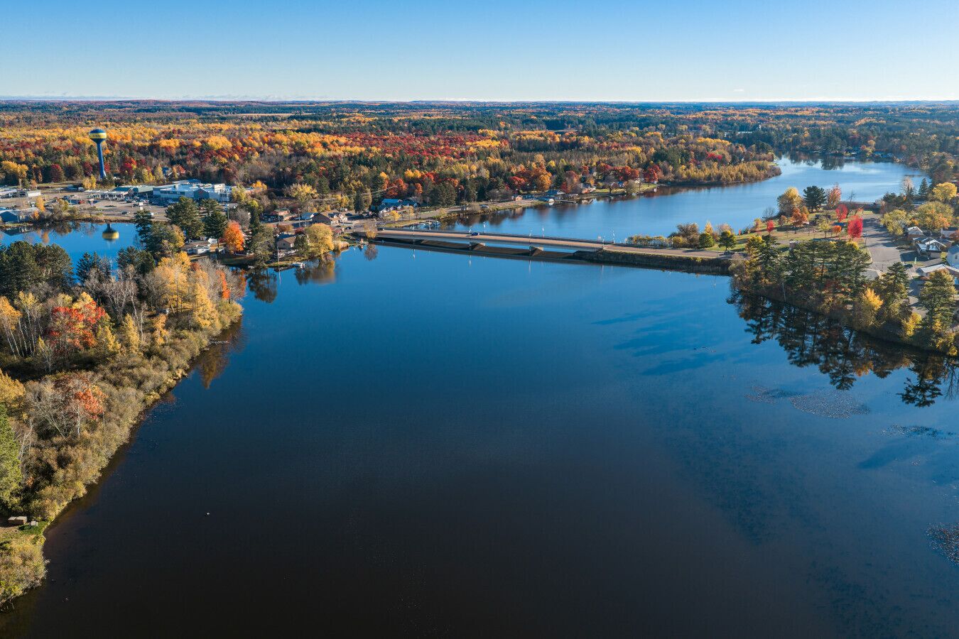 An aerial view of a lake surrounded by autumn-colored trees, a small dock, and a distant water tower.