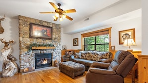 A cozy living room with a stone fireplace, a ceiling fan, and a large window. - 2BD + Loft, Walk-In Cabin- Discounts on Golf & Tickets! (Branson)