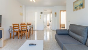 Un salon avec un canapé gris, un coin repas avec des chaises en bois et une table, et une cuisine à l’arrière-plan.