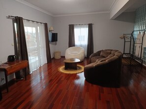 A living room with a wooden floor, a white armchair, a brown sofa, a coffee table, and a TV mounted on the wall. - Hôtel Les 12 Collines, Ivato Ambohijanahary (Antananarivo)