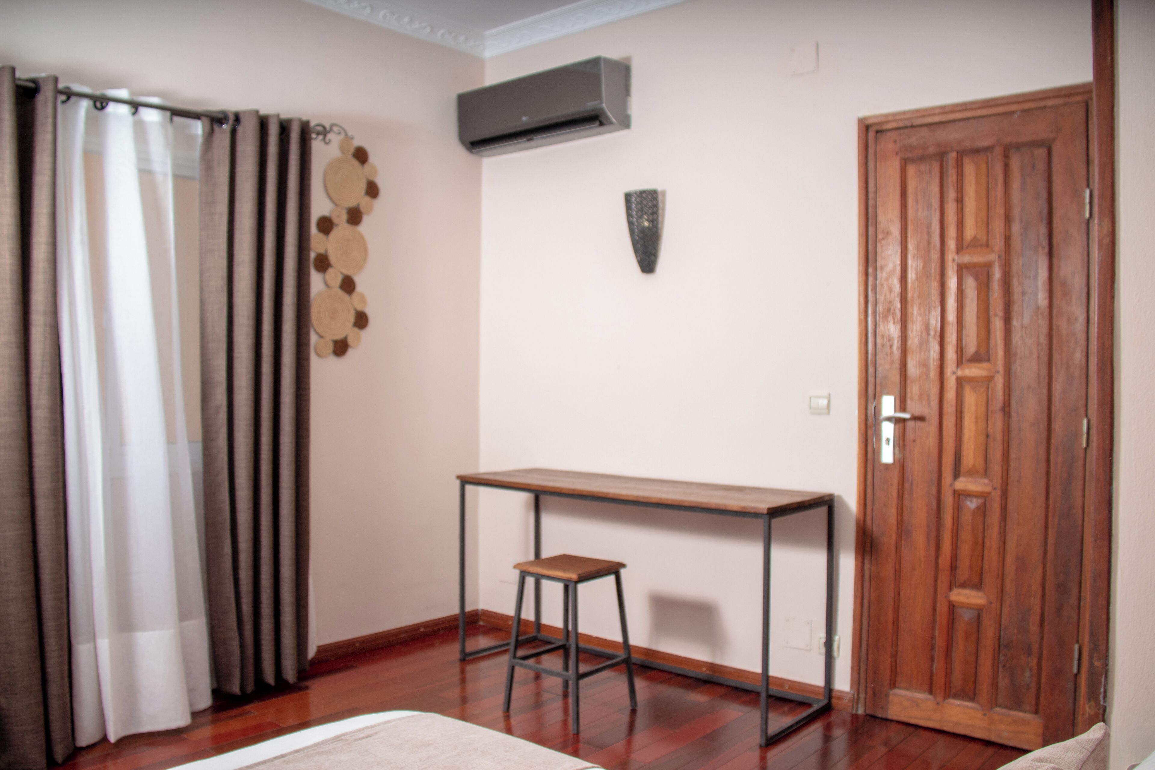 Une pièce avec un plancher en bois, une petite table en bois accompagnée d’un tabouret, une porte en bois et un rideau.
