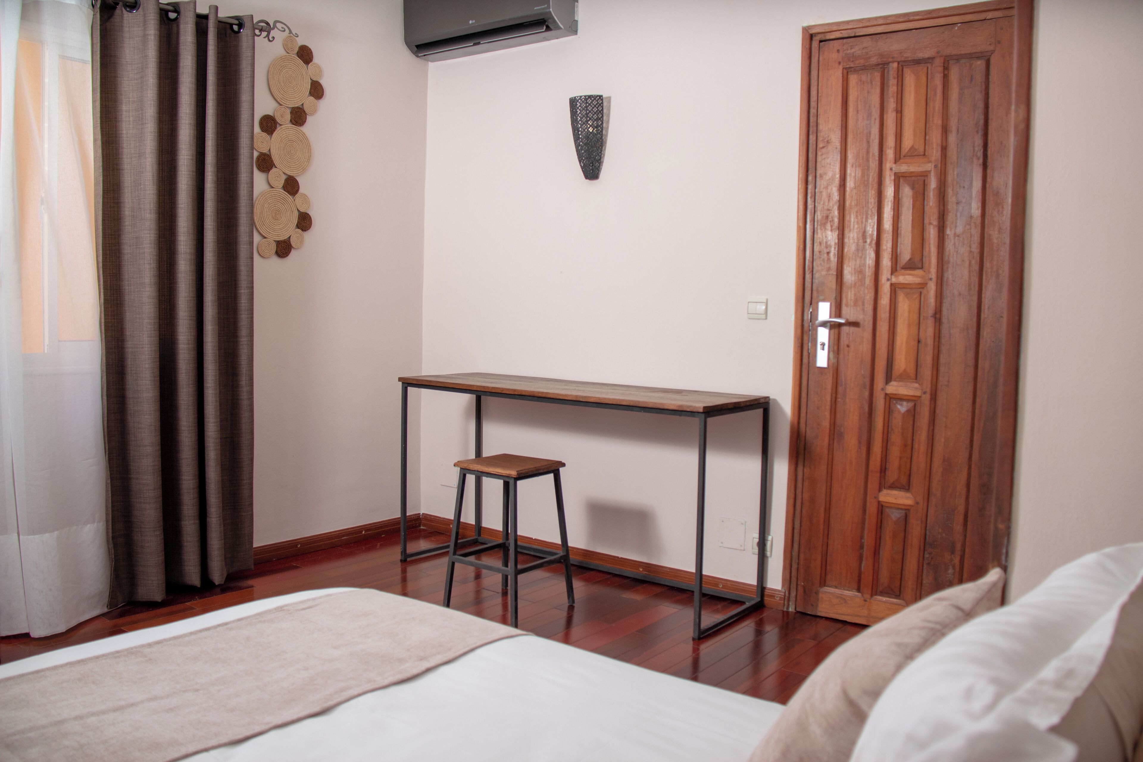 A room with a bed, a wooden desk with a stool, a wooden door, and a wall-mounted air conditioner.