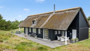 Een zwart houten huis met rieten dak, schoorsteen en een houten terras met buiten zitplaatsen.