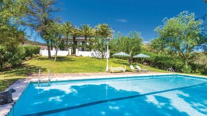 Una piscina con agua cristalina azul, rodeada de césped, árboles y un edificio blanco al fondo.