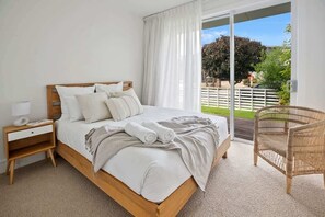 A bedroom with a wooden bed, white bedding, a nightstand, a wicker chair, and a view of a garden through a sliding door. - Casa Luxe (Kingscliff)