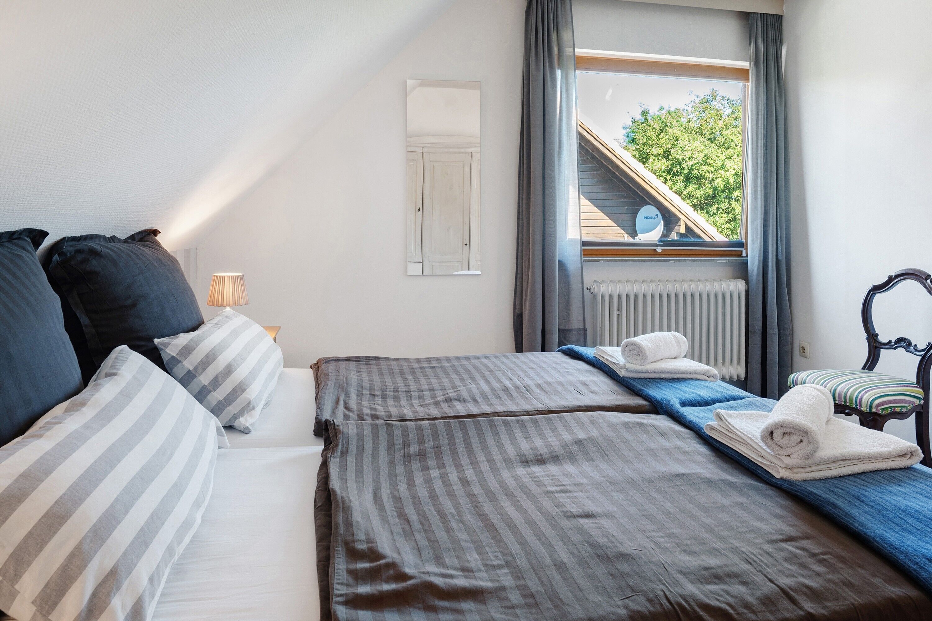 Een slaapkamer met een bed, kussens, een nachtkastje, een lamp, een raam met uitzicht op een gebouw en bomen, en een radiator.