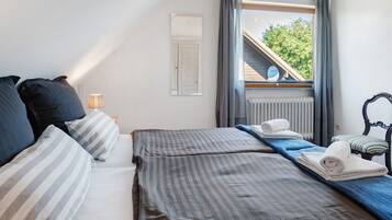 Een slaapkamer met een bed, kussens, een nachtkastje, een lamp, een raam met uitzicht op een gebouw en bomen, en een radiator.