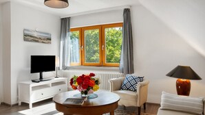 A living room with a TV, a round table with a book and flowers, a sofa, a lamp, and a window with curtains. - Apartment 'Ferienwohnung Lisa' with Private Terrace, Private Garden and Wi-Fi (Behrensdorf)