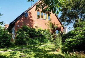 A brick house with a sloped roof, a visible satellite dish, and a garden with various plants and a pathway leading to the entrance. - Apartment 'Ferienwohnung Lisa' with Private Terrace, Private Garden and Wi-Fi (Behrensdorf)