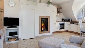 Ein modernes Wohnzimmer mit Flachbildfernseher, Kamin, Sofa und einer Küche mit weißen Schränken und Herd.