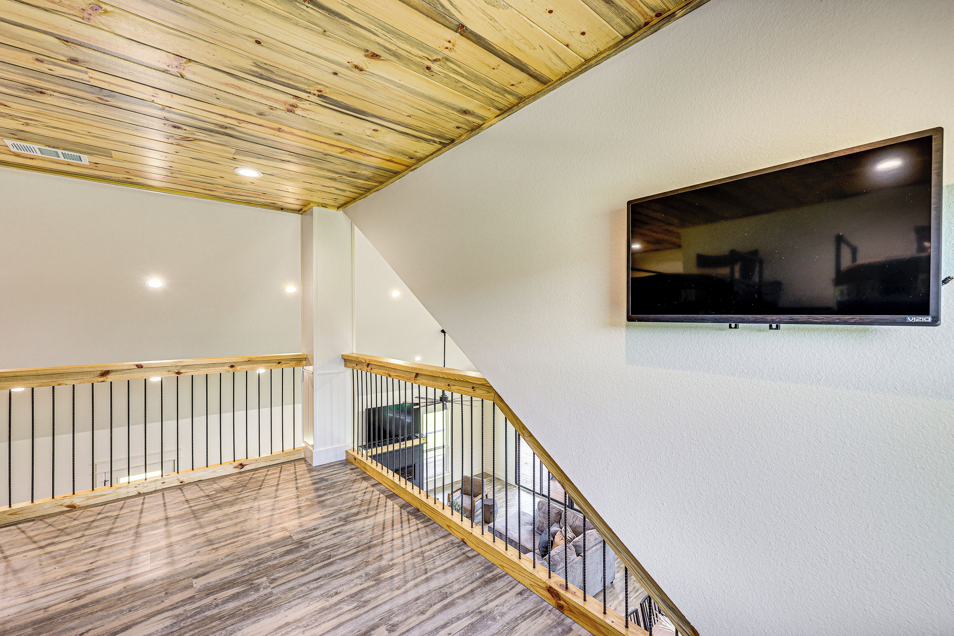 A spacious room with a wooden ceiling, a staircase with a wooden railing, and a flat-screen TV mounted on the wall.