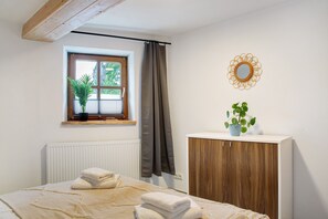Ein Schlafzimmer mit Bett, Fenster mit Pflanze, Heizkörper, Holzschrank und Spiegel an der Wand.